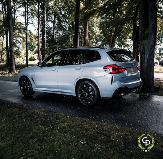 En elegant BMW X3 xDrive30e hybrid SUV med sporty sorte fælge er parkeret langs en skyggefuld kørebane, omgivet af frodige grønne træer, der viser en blanding af luksus og natur.