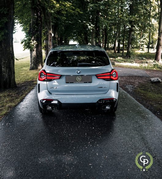 En sølvfarvet BMW X3 xDrive30e plug-in hybrid parkeret på en våd vej foret med træer, med karakteristiske LED-baglygter, der lyser, lige efter en regnbyge.