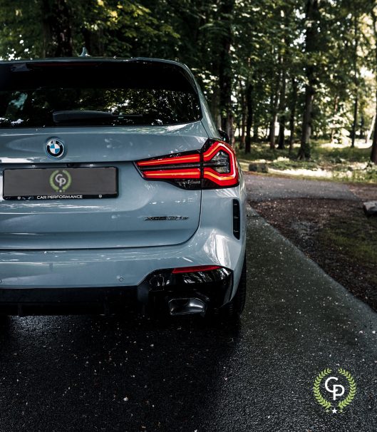 En BMW X3 xDrive30e hybrid SUV med karakteristiske baglygter parkerer på en våd vej midt i frodige grønne områder og viser sit sporty design og "m performance"-mærket, der antyder