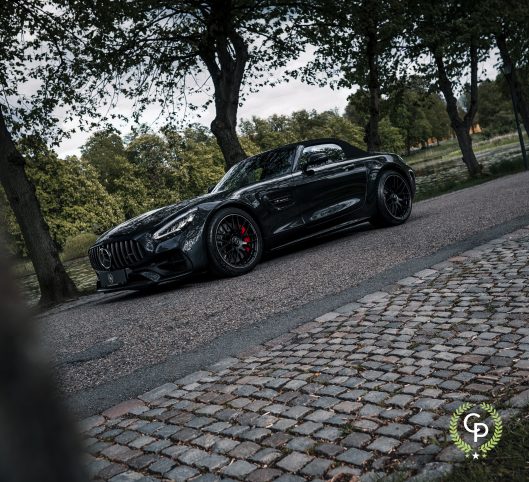 En elegant sort Mercedes-AMG GT Roadster parkeret på en brostensbelagt sti, der emmer af luksus og kraft, omgivet af fredfyldte parkgrønt under en overskyet himmel.