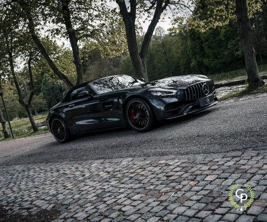 En slank sort Mercedes AMG GT C Roadster parkeret på en brostensbelagt sti, der viser sit dynamiske design og sportsvognstiltrækning midt i rolige parkomgivelser.