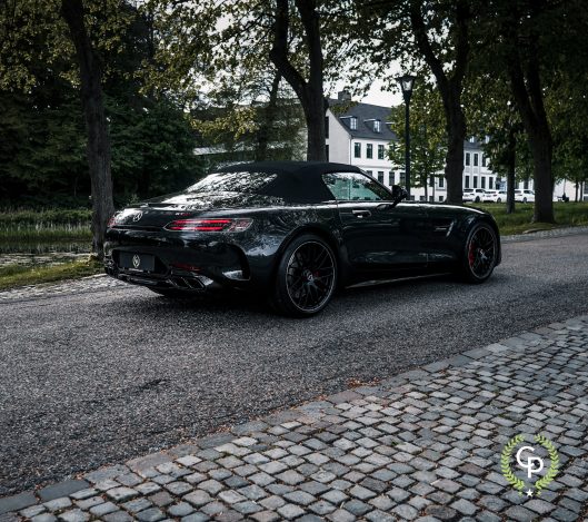 En slank sort Mercedes AMG GT cabriolet sportsvogn parkeret på en brostensbelagt sti, der praler med sit aggressive design og luksushjul, omgivet af rolig parklandskab med træer.