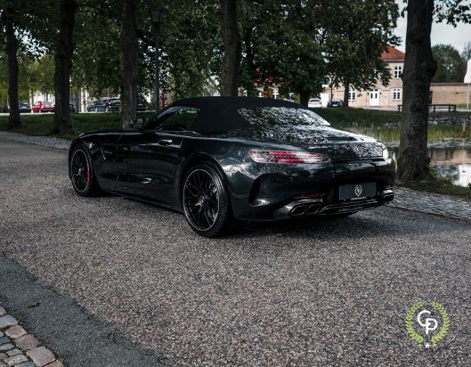 En elegant sort Mercedes AMG GT C Roadster parkeret på en brostensbelagt gade ved siden af en rolig vandvej med træer og en klassisk bygning i europæisk stil i baggrunden.