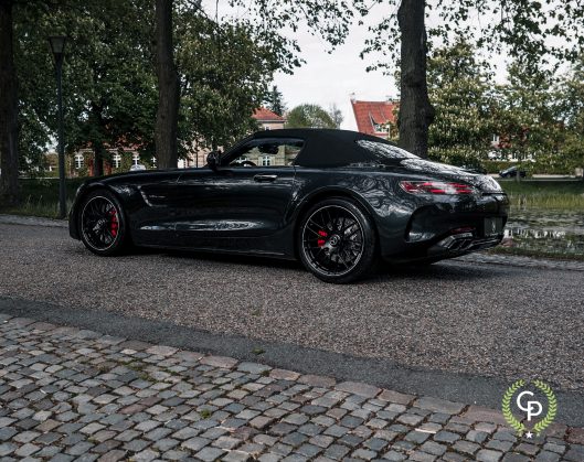 En elegant sort Mercedes AMG GT roadster med røde bremsekalipre parkeret på en brostensbelagt vej, omgivet af frodige grønne træer og traditionel europæisk arkitektur.