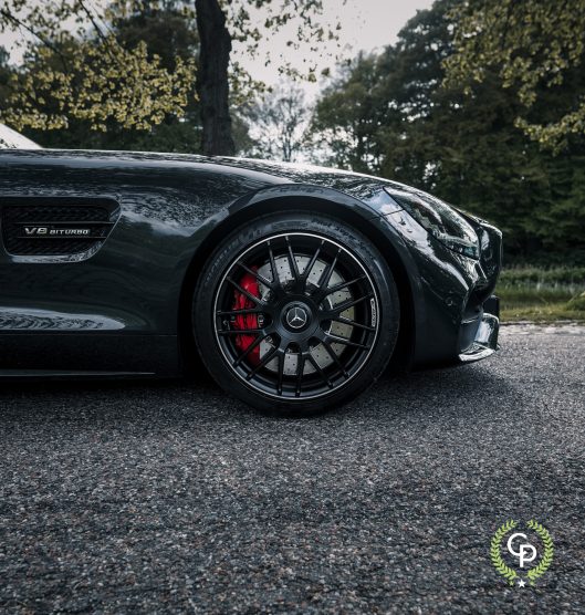 Et nærbillede af en sort Mercedes AMG GT Roadster V8 biturbo, med fokus på dens blanke maling, stilfulde alufælge med karakteristiske bremsekaliber, optaget på en rolig baggrund af