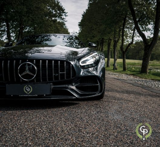 En elegant sort Mercedes Amg GT Roadster parkeret på en brostensbelagt sti og fremviser sin kraftfulde frontgrill og emblem under en baldakin af træer, der emmer af luksus og ydeevne.