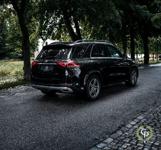 En slank sort Mercedes GLE350de parkeret på en brostensbelagt gade ved siden af en fredfyldt park og viser dens blanke finish og moderne design under en baldakin af frodige træer på en overskyet dag.