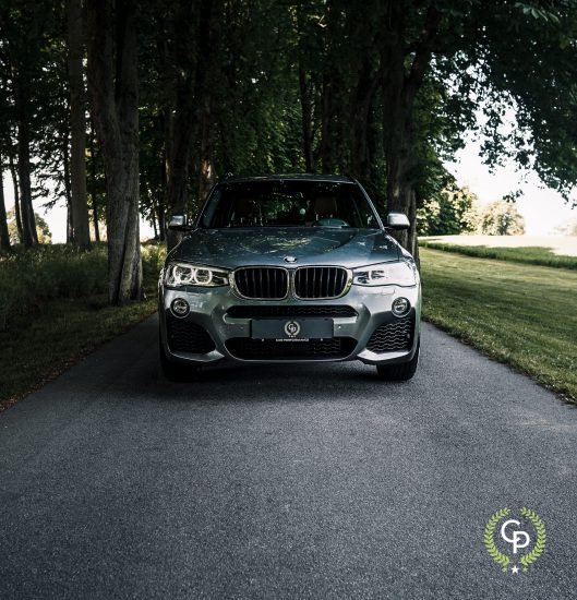 En elegant sølv BMW X3, en luksus-SUV, parkerer på en skyggefuld vej, flankeret af frodige grønne træer under en klar himmel, og udstråler essensen af tysk bilhåndværk og løftet om