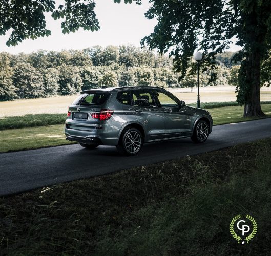 En sølvfarvet BMW X3 parkeret på en fredfyldt, træomkranset vej, der viser sit slanke design og sporty silhuet mod den grønne kulisse af en velholdt græsklædt park under en klar blå himmel