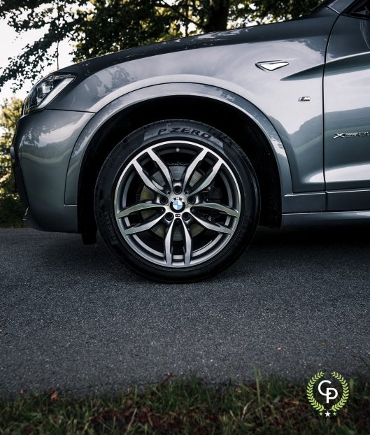 Et nærbillede af en BMW X3's forhjul, der viser dens stilfulde alufælge og Pirelli P Zero-dæk, delvist oplyst af naturligt lys på baggrund af udendørs.