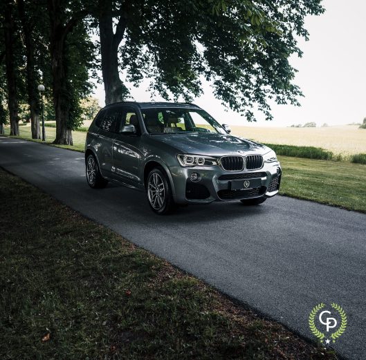 En slank grå BMW X3 SUV parkeret på en idyllisk træklædt vej, der sammenstiller luksuskøretøjets moderne teknik mod en rolig, naturlig baggrund.