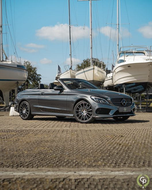 En stilfuld Mercedes C43 Cabriolet parkerer selvsikkert blandt forankrede yachts og inkarnerer luksus og essensen af fritid under en klar blå himmel.