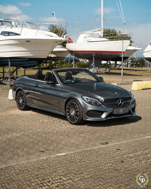 En slank, mørkegrå Mercedes C43 Cabriolet parkeret ved en lystbådehavn med en baggrund af både på tørdokker under en klar blå himmel, der viser luksus til lands og til vands.