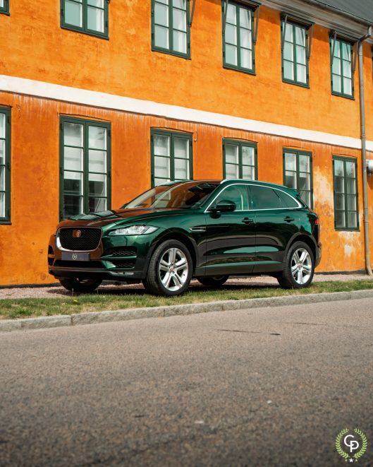 En elegant grøn Jaguar F-Pace, en luksus-SUV, parkeret på en brostensbelagt gade ved siden af en livlig orange bygning, der emmer af ydeevne og moderne design i historiske bymiljøer.