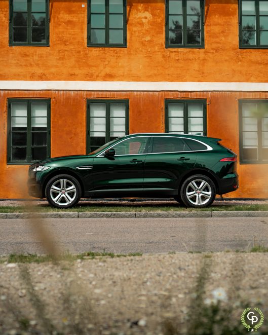 En slank grøn Jaguar F-Pace, en luksus SUV, parkeret på en gade, med dens polerede overflade, der afspejler den orange facade af en vintage bygning, der legemliggør en blanding af moderne mobilitet og gammeldags